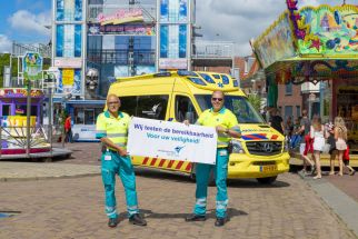 Foto van twee ambulancemedewerkers die een bord vasthouden waarop staat: Wij testen de bereikbaarheid voor uw veiligheid. Op de achtergrond staat een geparkeerde ambulance midden op een kermisterrein. 