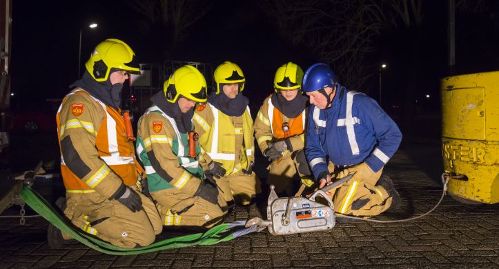 Foto van vier brandweermensen in opleiding en een instructeur die in het donker op de grond bij een trekhaak zitten.