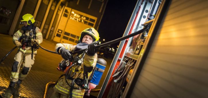 Foto van twee brandweermensen in opleiding die een oefening doen met een brandslang naast een brandweerwagen.
