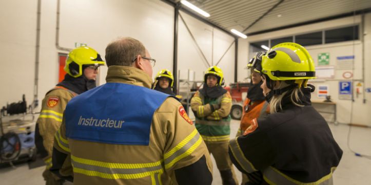 Foto van een instructeur en een groepje brandweermensen in opleiding die in een kring staan tijdens een uitleg. 