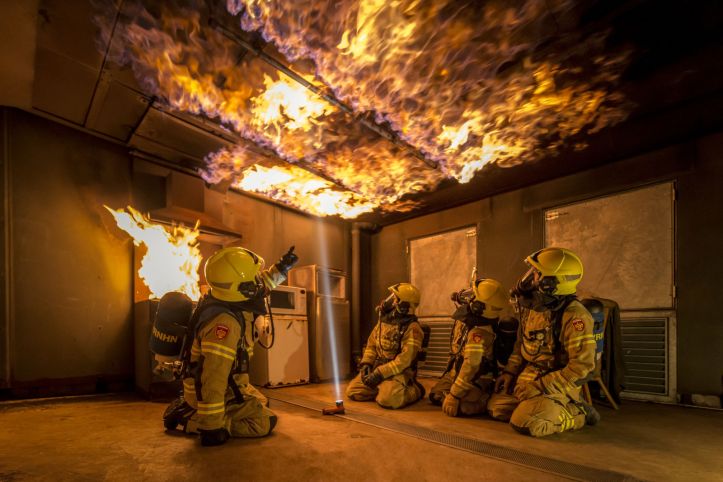 Foto van een instructeur en drie brandweermensen in opleiding in een oefenruimte waarbij het plafond in brand staat.