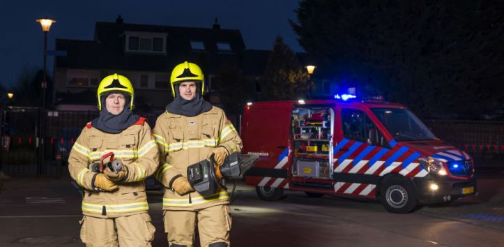 Foto van twee brandweermensen met op de achtergrond een SI2 voertuig met brandende zwaailichten. 